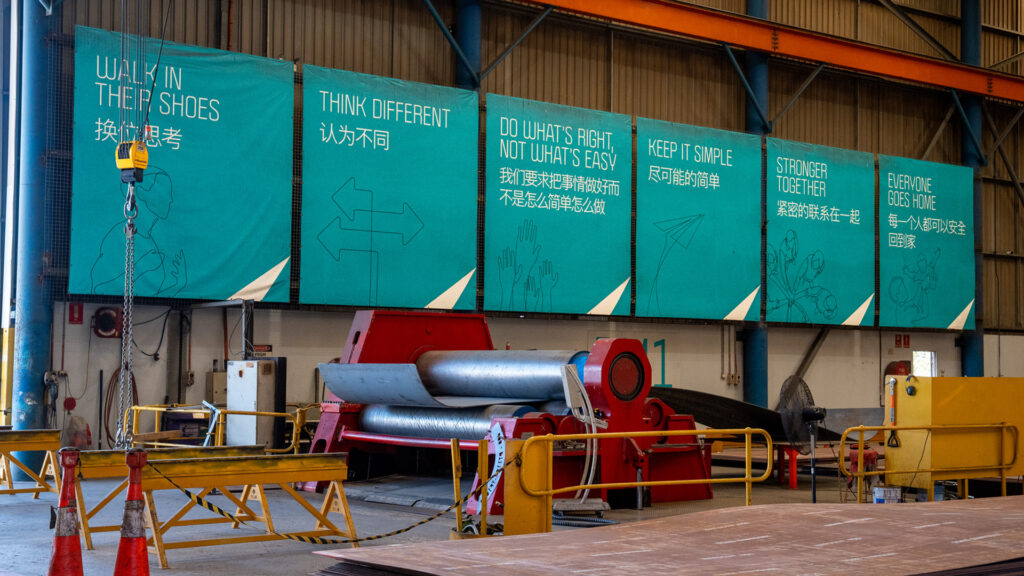 Photo of the multilingual signs in Schlam's Forrestfield. Signs read: WALK IN THEIR SHOES, THINK DIFFERENT, DO WHAT'S RIGHT NOT WHAT'S EASY, KEEP IT SIMPLE, STRONGER TOGETHER, EVERYONE GOES HOME. A sheet metal roller is in front of the signs.